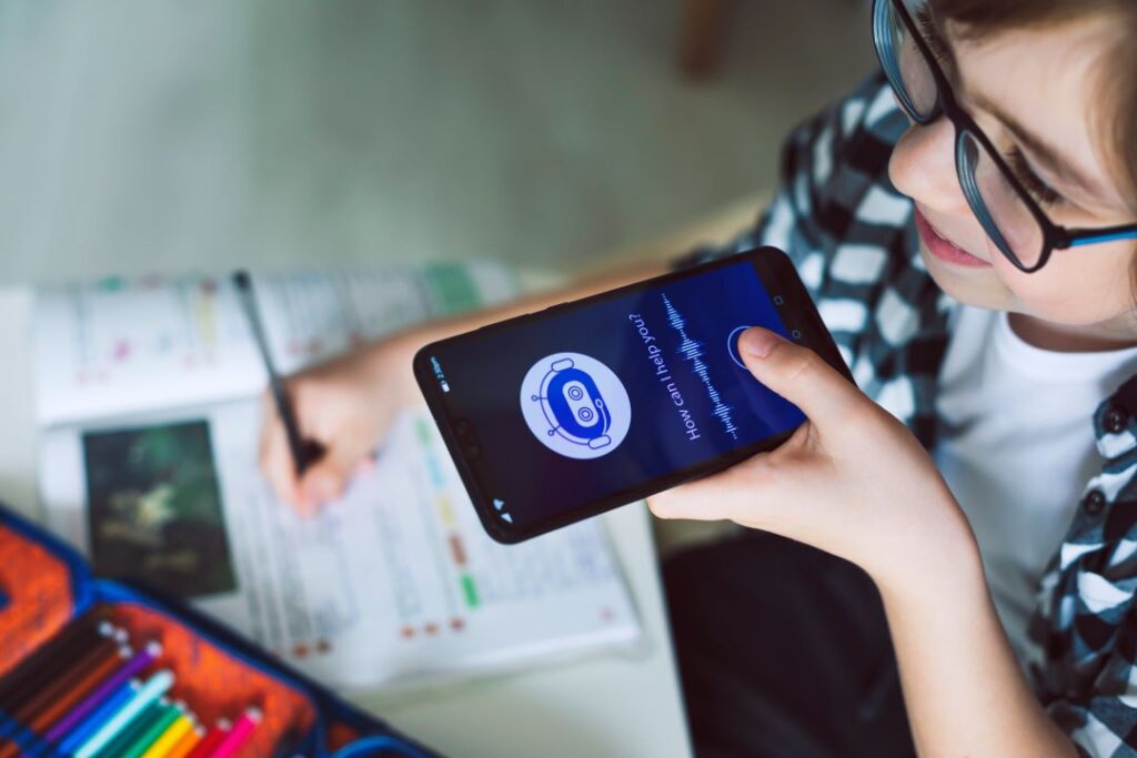 Imagem de uma mulher branca falando para a plataforma de inteligência artificial no celular. Ela está estudando, então, em cima da mesa tem cadernos e canetas.