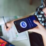 Imagem de uma mulher branca falando para a plataforma de inteligência artificial no celular. Ela está estudando, então, em cima da mesa tem cadernos e canetas.