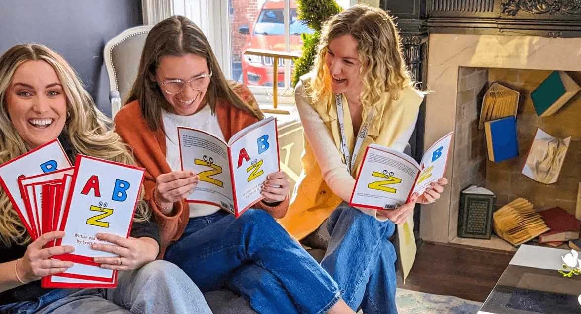 Três senhoras, da equipe WebFX, sentadas no sofá compartilhando o livro infantil AB a Z, sorrindo e rindo em uma sala decorada com livros