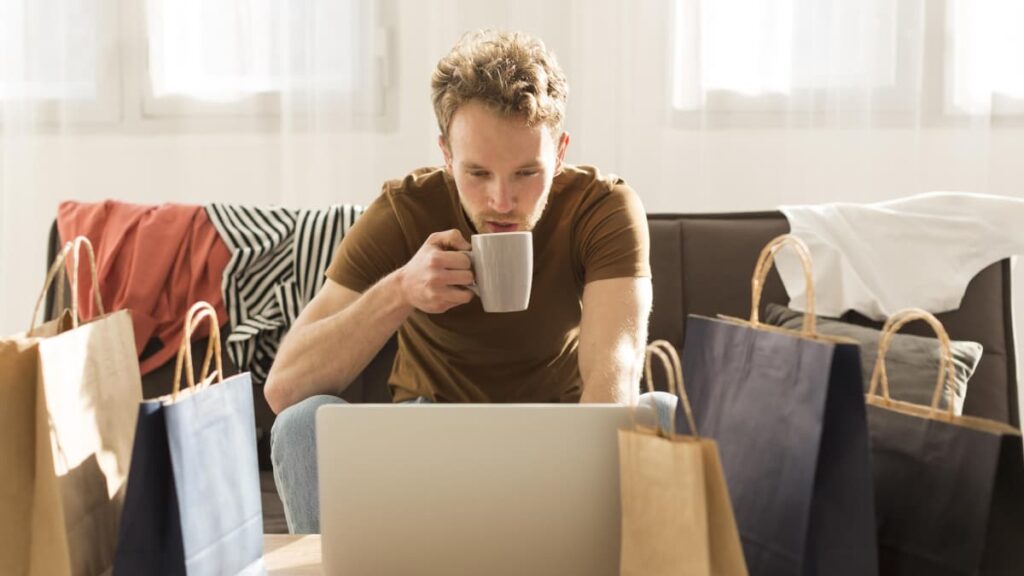 Rapaz em frente ao computador, com sacos de papel à sua volta (a representar compras online). Está a segurar uma chávena