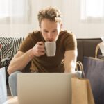 Rapaz em frente ao computador, com sacos de papel à sua volta (a representar compras online). Está a segurar uma chávena