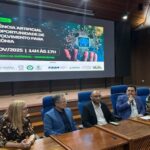 Foto mostra homem de blazer azul falando ao microfone em um evento